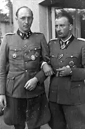 A black-and-white photograph of two men wearing military uniforms with various military decorations attached to their uniforms. The man on the left is wearing glasses and is smoking a cigarette, the man on the right is smoking a cigar.