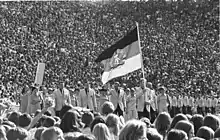 People march behind a large flag surrounded by spectators