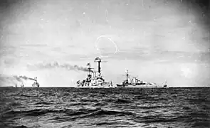 Two large warships steaming in opposite directions close together, with several other vessels are in the distance.