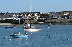 Foreground: A handful of tethered boats floating on calm water, surrounded by small buoys. Background: Raised shoreline of rock and concrete, topped by boats lined up beside each other; in the centre a slipway meets the water. In the far background is a collection of small residential buildings.