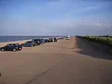 cars lined up stretching away along hard surfaced standing next to a golden sand beach and sea beyond against a blue sky on a sunny day