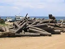 An unorganized pile of approximately 50 rosewood logs sits on a beach with boats in the background.