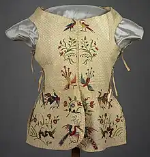A loose vest-like garment in a faded cream color with bright red and green embroidery depicting plants and birds.