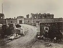 Photograph of the fort of Vanvres, destroyed by the war. In the foreground, a road leading up to it, ending in a bridge. It crosses a low wall which is partly destroyed. In the second plan, the fort's wall and an entrance, located behind a ditch and only slightly damaged. Behind the wall, a few buildings can be seen, including a large barracks of which only parts of the facades remain.