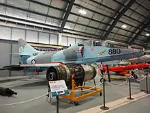 Colour photograph of a military fighter jet aircraft painted in blue and grey camouflage in a museum, with other aircraft and engines visible