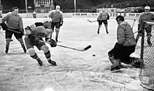 Black and white in-game action photo of a United States player shooting the puck at the net surrounded by Yugoslavia players