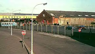 Fenced-off tarmac car parking area, with a run-down red-brick industrial building in the background.