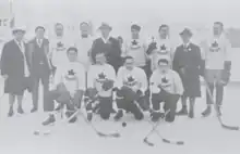 Black and white photo of a hockey team outside on natural ice, including four men each dressed in a dark overcoat, and nine players dressed in hockey equipment white sweaters with a maple leaf crest