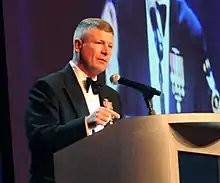 MCPON Rick D. West standing at a lectern with a projection screen and two flags behind him.